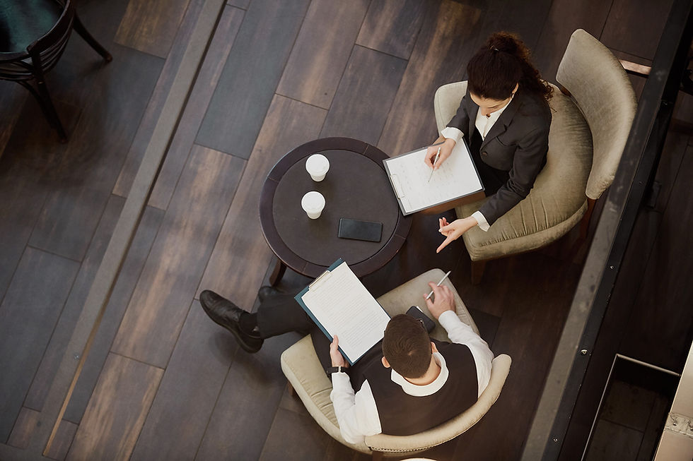 Two people in business attire sit at a round table with notebooks in a wood-floored setting. Coffee cups and a phone are on the table.