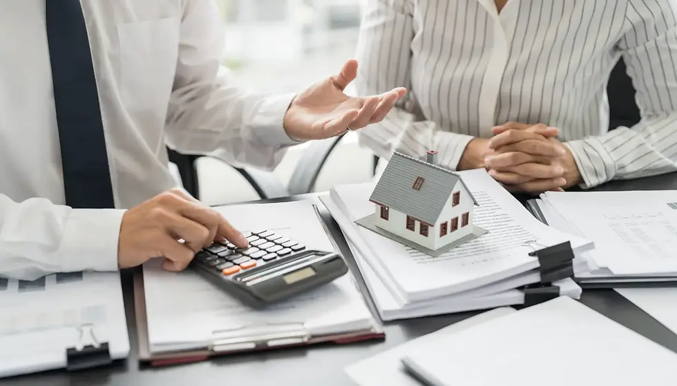 Two people discussing building insurance with papers and a house model on a table. One uses a calculator, the other listens attentively, suggesting focus.