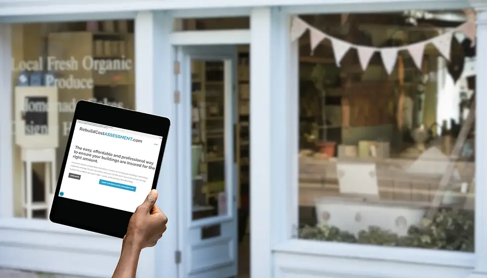 Hand holding a tablet displaying RebuildCostAssessment.com, in front of a shop with a pastel blue exterior. A banner decorates the storefront.