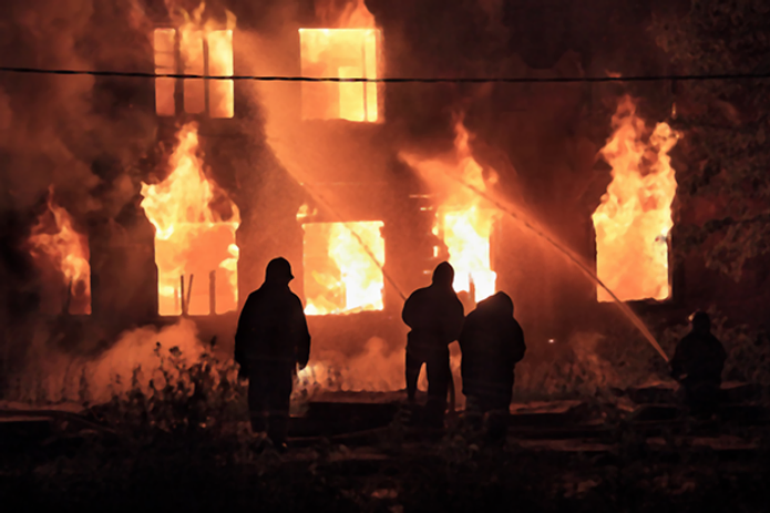 Silhouetted firefighters combat a blazing two-story building fire at night. Intense flames and smoke fill the dark sky, creating urgency.