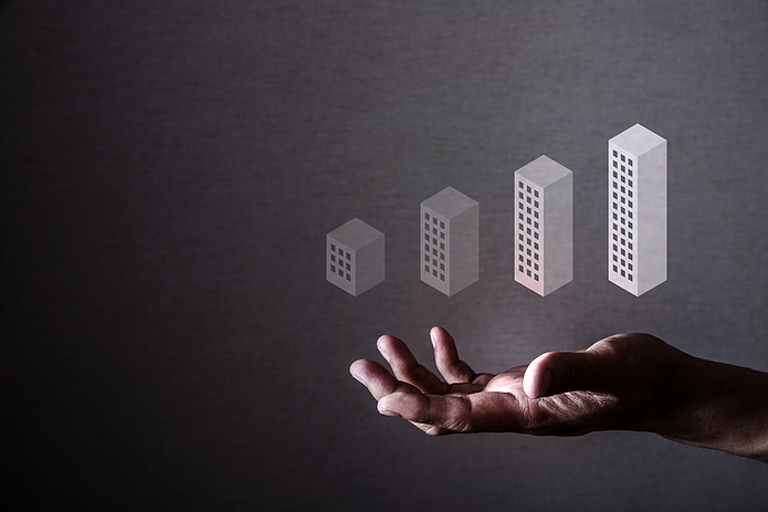 A hand under four floating 3D graph bars resembling buildings, in a dark setting. The bars increment in height, suggesting growth.