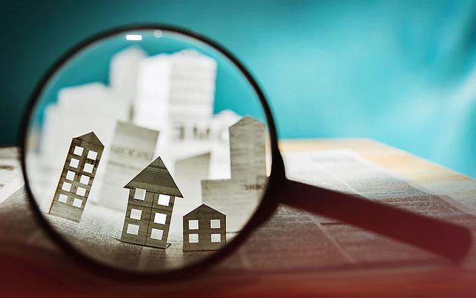 Magnifying glass focuses on paper cut-out buildings, on a newspaper backdrop. Bright teal background adds contrast.